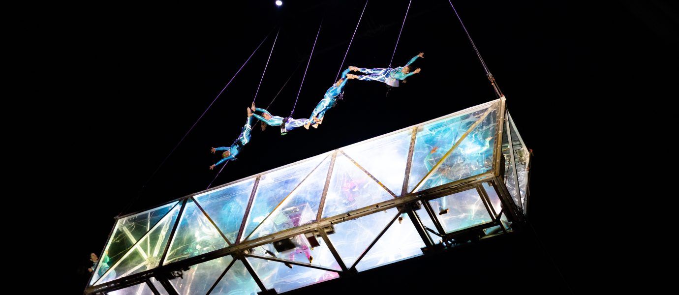 Five acrobats perform an aerial routine above a transparent, illuminated glass structure suspended in the air, with black background and stage lights shining from above.