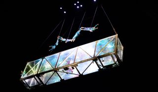 Five acrobats perform an aerial routine above a transparent, illuminated glass structure suspended in the air, with black background and stage lights shining from above.