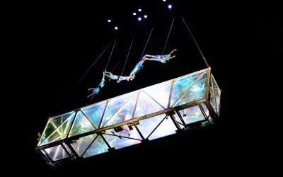 Five acrobats perform an aerial routine above a transparent, illuminated glass structure suspended in the air, with black background and stage lights shining from above.