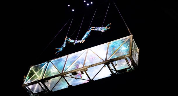 Five acrobats perform an aerial routine above a transparent, illuminated glass structure suspended in the air, with black background and stage lights shining from above.