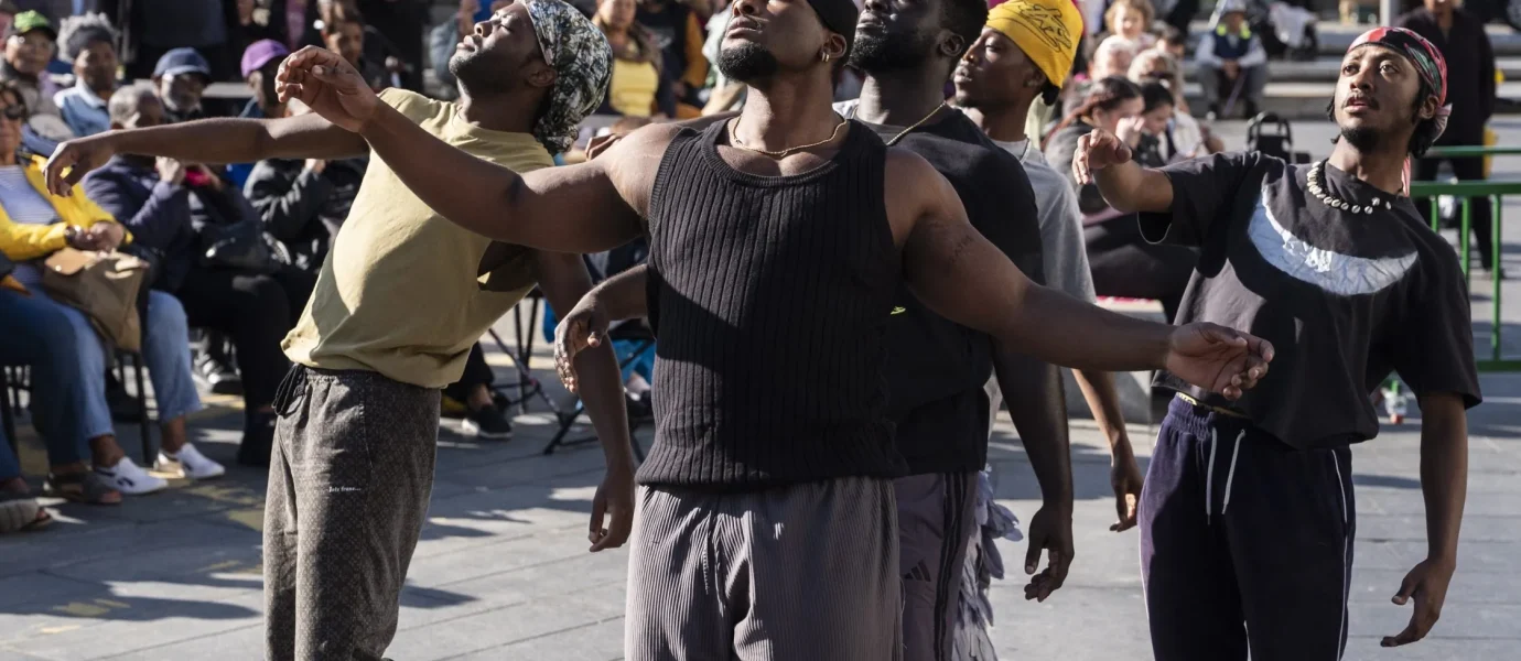 Five men perform an expressive contemporary dance outdoors in a city square, with a crowd of people watching in the background. The dancers wear casual clothing and appear deeply focused on their movements.