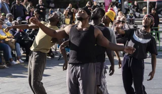 Five men perform an expressive contemporary dance outdoors in a city square, with a crowd of people watching in the background. The dancers wear casual clothing and appear deeply focused on their movements.