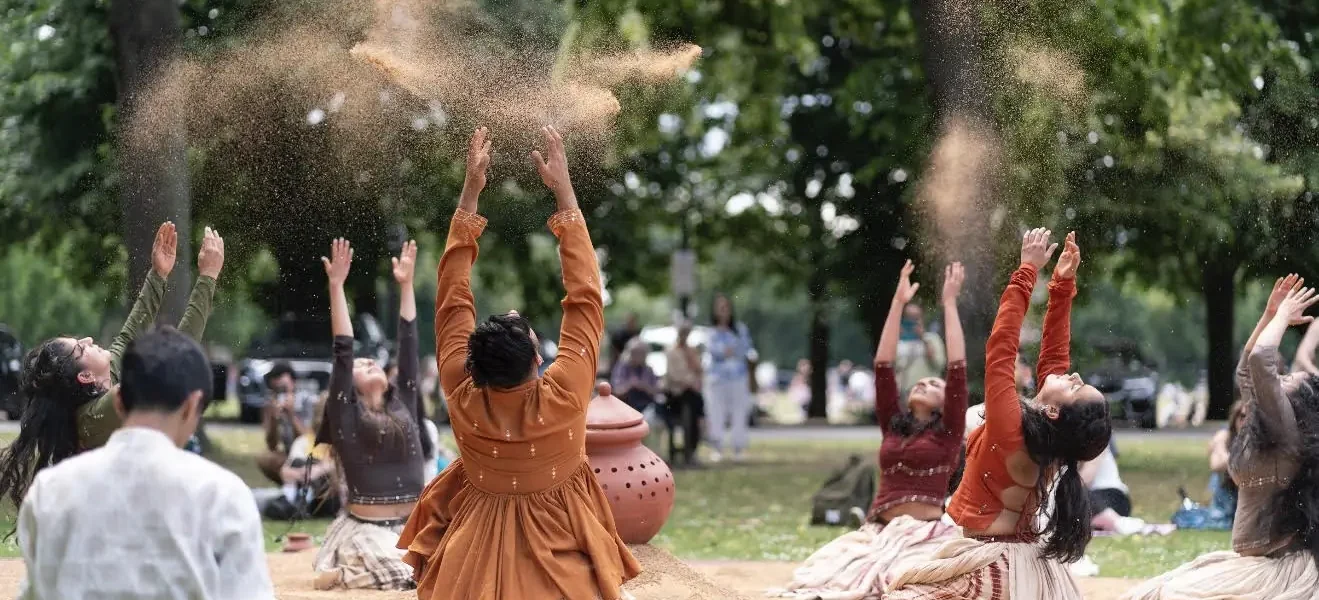 A group of people in earthy-toned clothing sit on the ground in a park, raising their arms and tossing sand into the air, surrounded by trees and other people in the background.