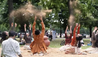 A group of people in earthy-toned clothing sit on the ground in a park, raising their arms and tossing sand into the air, surrounded by trees and other people in the background.