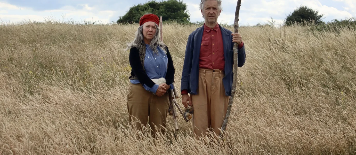 Two older adults stand side by side in a tall grassy field. The woman wears a red headband, blue shirt, and brown trousers; the man holds a tall stick and wears a blue jacket, red shirt, and brown trousers. Bushes and sky are in the background.