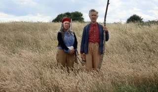 Two older adults stand side by side in a tall grassy field. The woman wears a red headband, blue shirt, and brown trousers; the man holds a tall stick and wears a blue jacket, red shirt, and brown trousers. Bushes and sky are in the background.