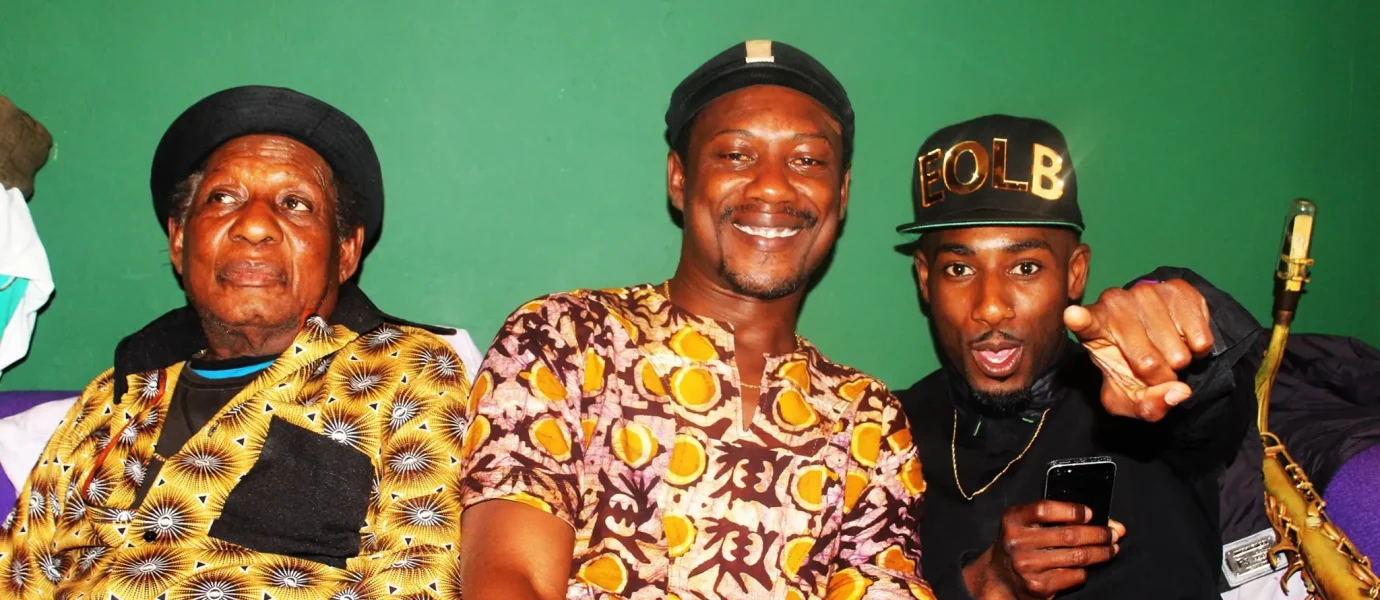 Three men sitting on a sofa against a green wall. The man on the left wears a patterned outfit and cap, the man in the middle wears a patterned shirt and smiles, and the man on the right wears a black outfit and points forward.