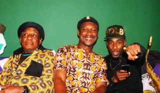Three men sitting on a sofa against a green wall. The man on the left wears a patterned outfit and cap, the man in the middle wears a patterned shirt and smiles, and the man on the right wears a black outfit and points forward.