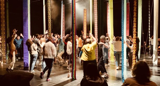 A group of people dance energetically among tall, colorful hanging ribbons on a dimly lit stage, while others sit and watch from the edges.