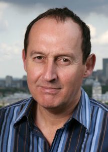 A middle-aged man with short dark hair wears a blue striped shirt and stands outdoors with a neutral expression. The blurred background shows a cityscape under a cloudy sky.