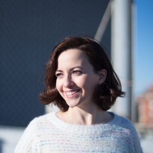 A woman with short brown hair, wearing a white sweater with pastel stripes, smiles while standing outdoors in bright sunlight, with a blurred urban background behind her.