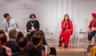 Five people sit on stage in front of an audience, engaged in a panel discussion. One woman in a red suit speaks into a microphone while the others listen. The setting appears to be a conference or seminar.