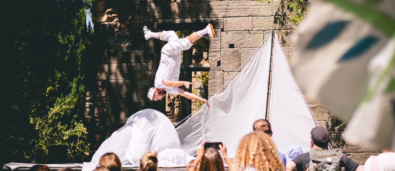 A performer dressed in white does a handstand on stage outdoors in front of an audience. The stage has white tents and is set against a stone wall, with trees and people watching and taking photos in the foreground.