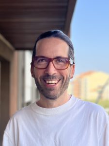 A smiling man with long dark hair tied back, wearing large brown glasses, a white shirt, and small earrings, stands outside with blurry buildings and a blue sky in the background.