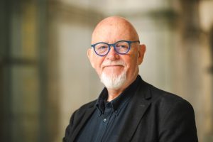 An older man with a bald head and white goatee wearing blue glasses, a dark shirt, and a black jacket, stands smiling in front of a blurred, light-colored background.