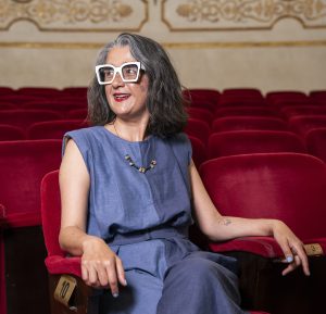 A woman with gray hair, wearing large white glasses and a blue outfit, sits smiling in a red velvet theater seat with ornate decor in the background.
