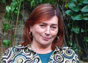A woman with shoulder-length auburn hair and gold hoop earrings stands outdoors in front of green leafy plants, wearing a patterned shirt and giving a gentle, knowing smile.