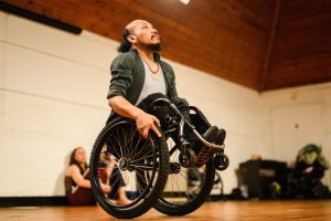 A man in a wheelchair lifts himself using the wheels in a spacious room with wooden ceilings. In the background, a seated woman watches him. The scene suggests an indoor activity or rehearsal.