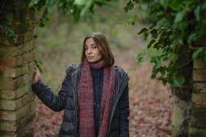 A woman in a dark coat and red scarf stands between two old brick pillars covered with green ivy, in a forest with fallen leaves on the ground, looking calmly at the camera.