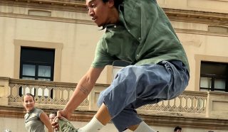 A dancer leaps mid-air during an outdoor performance, one arm raised and legs bent. A crowd watches in the background, with a woman smiling nearby. The setting is a sunny day beside a beige building.