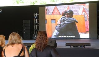 An outdoor film screening showing two men dressed in black in an embrace in an urban setting