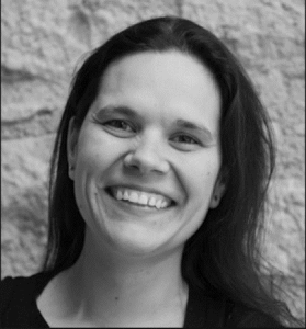 A woman with long dark hair smiles warmly at the camera. She is wearing a dark top and standing in front of a textured stone wall. The image is in black and white.
