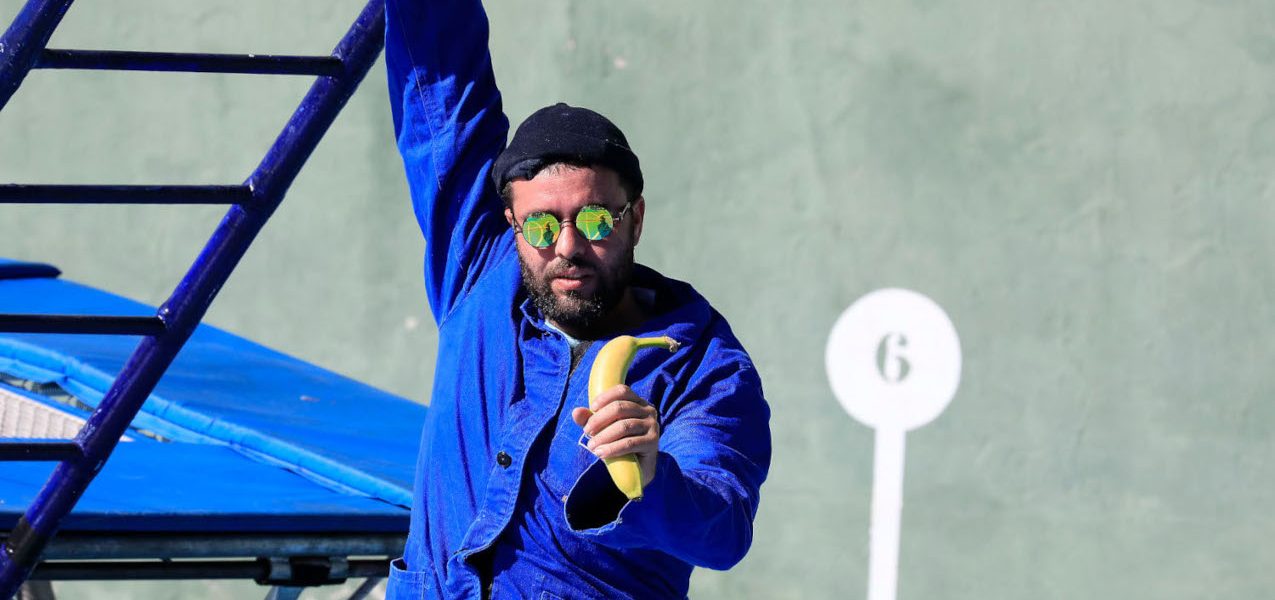 A man in a blue jacket and sunglasses holds a banana and hangs onto a blue metal structure. He stands in front of a green wall marked with a white circle and the number 6.