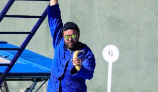 A man in a blue jacket and sunglasses holds a banana and hangs onto a blue metal structure. He stands in front of a green wall marked with a white circle and the number 6.