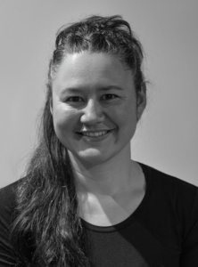 A woman with long, dark hair, partly tied back, smiles at the camera. She is wearing a dark, crew-neck top and is photographed against a plain, light background in black and white.