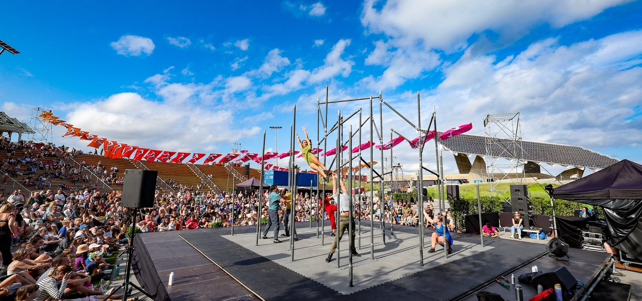 Performers on a stage with metal bars entertain a large outdoor audience under a bright blue sky with scattered clouds. Colorful banners and stadium seating are visible in the background.