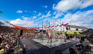 Performers on a stage with metal bars entertain a large outdoor audience under a bright blue sky with scattered clouds. Colorful banners and stadium seating are visible in the background.