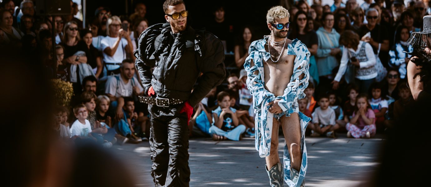 Two models walk confidently down an outdoor catwalk, surrounded by a seated crowd. One wears a black suit and sunglasses, the other dons a patterned blue coat, boots, sunglasses, and blue beads, with much of his chest and legs exposed.