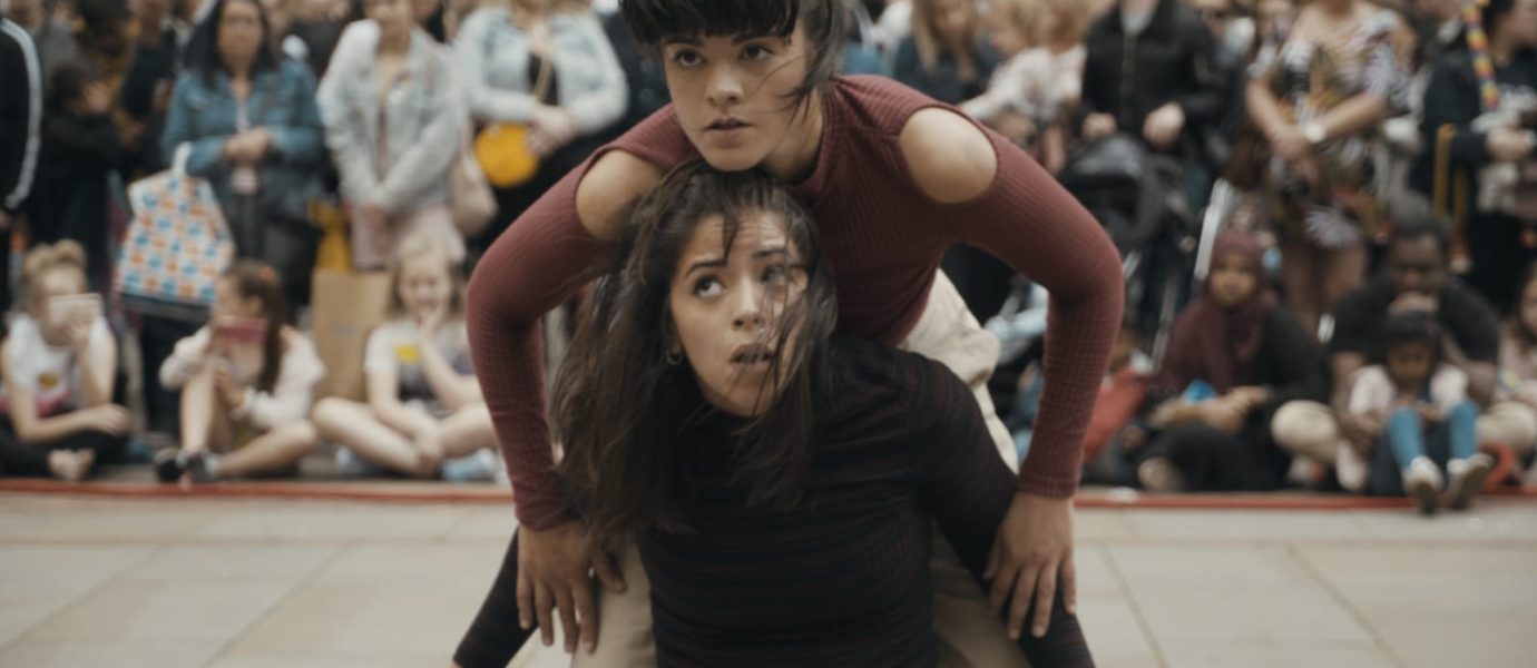 Two women perform an expressive dance routine outdoors, with one crouched beneath the other’s arms; a large, diverse crowd watches intently in the background.