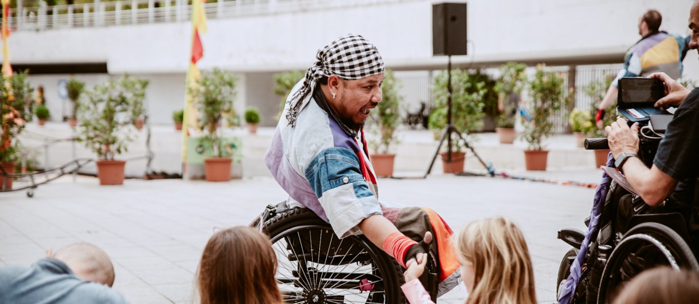A man in a wheelchair wearing a bandana and colourful clothing smiles and interacts with two young girls outdoors. Other people and potted plants are visible in the background.