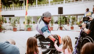 A man in a wheelchair wearing a bandana and colourful clothing smiles and interacts with two young girls outdoors. Other people and potted plants are visible in the background.