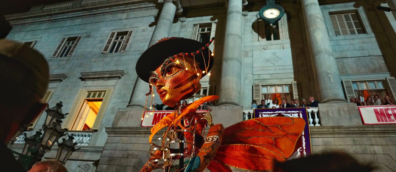 A large, colourful puppet with illuminated lights is displayed in front of a historic building at night. The puppet has orange wings and a hat, whilst people watch the scene from below and from balconies above.