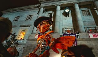 A large, colourful puppet with illuminated lights is displayed in front of a historic building at night. The puppet has orange wings and a hat, whilst people watch the scene from below and from balconies above.