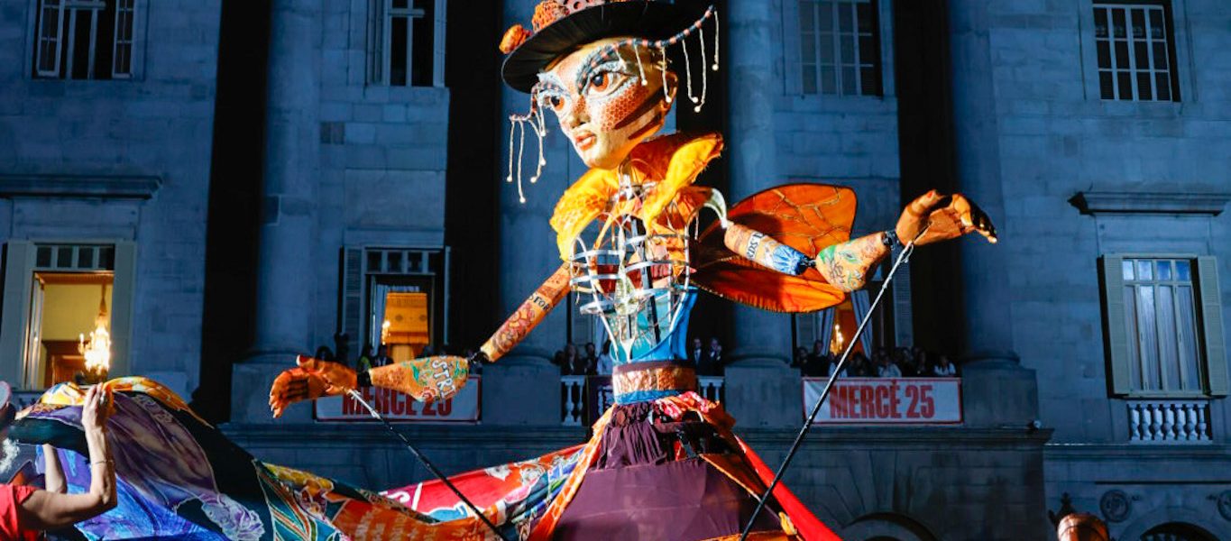 A large, colourful puppet with extended arms and a vibrant dress is paraded by performers at night in front of an historic building, as a crowd watches the lively street festival.