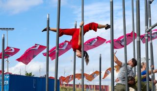 A performer in red trousers hangs upside down from a tall metal pole, balancing by her legs, while two people assist nearby. Colourful flags and a large crowd are visible in the background under a bright blue sky.