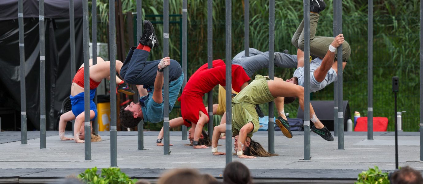 A group of performers does synchronised acrobatics, balancing upside down on parallel metal bars on an outdoor stage, as an audience watches. Each performer wears colourful clothing.