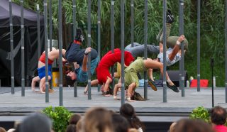 A group of performers does synchronised acrobatics, balancing upside down on parallel metal bars on an outdoor stage, as an audience watches. Each performer wears colourful clothing.