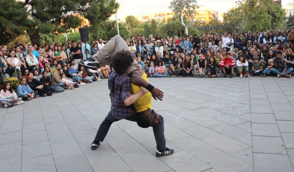 Two performers are dancing in the centre of a plaza, with one person lifting the other upside down. A large crowd sits and stands in a circle around them, watching the performance outdoors.