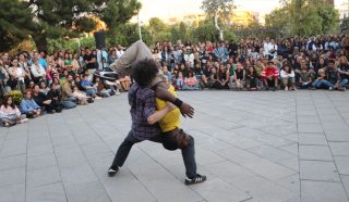 Two performers are dancing in the centre of a plaza, with one person lifting the other upside down. A large crowd sits and stands in a circle around them, watching the performance outdoors.