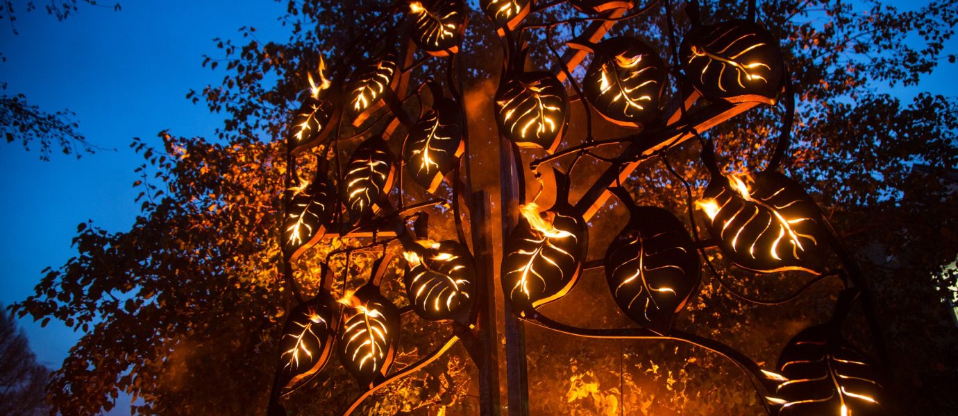 A metal sculpture shaped like a tree with leaf-shaped lanterns glows with fire against a twilight sky, surrounded by trees and soft orange light.