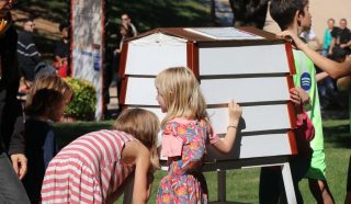 Three children closely examine a small, white, house-shaped structure outdoors while adults and other children are nearby on a sunny day. Some people are seated in the background on grass.