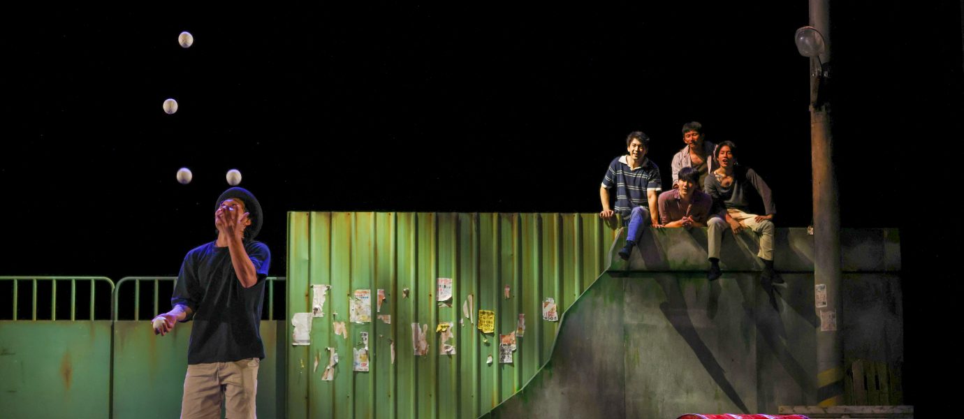 A performer in a hat juggles five balls on stage beside a red barrel, while four people sit on a raised ledge in the background, watching. The set resembles an urban street scene with a green metal fence.