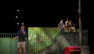 A performer in a hat juggles five balls on stage beside a red barrel, while four people sit on a raised ledge in the background, watching. The set resembles an urban street scene with a green metal fence.
