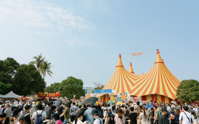 A large crowd gathers outdoors in front of a colourful yellow and orange striped circus tent with pointed tops, surrounded by trees under a bright, partly cloudy sky.