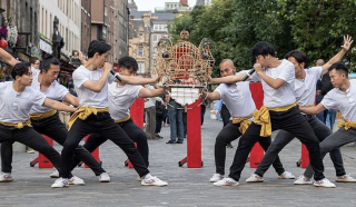 A group of nine men in matching white shirts and yellow sashes perform synchronised martial arts moves outdoors on a city street, with a decorative structure and trees in the background.