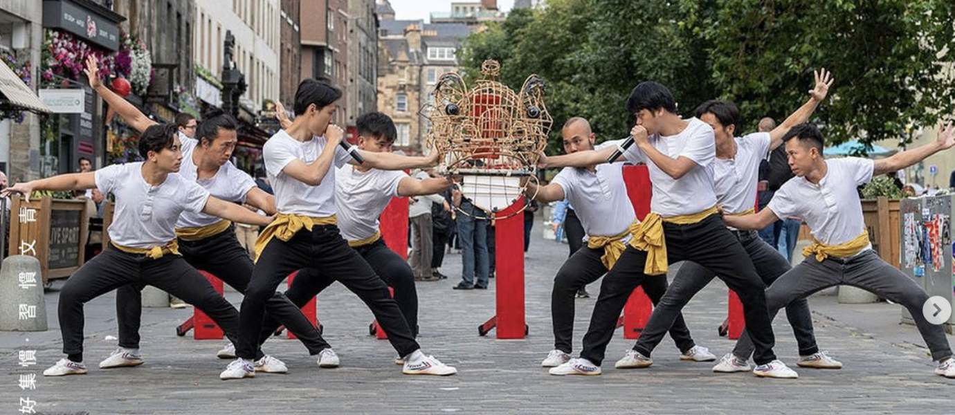 A group of nine people in white shirts and black trousers perform a synchronised martial arts pose on a cobblestone street, with red pillars and a decorative structure in the background.